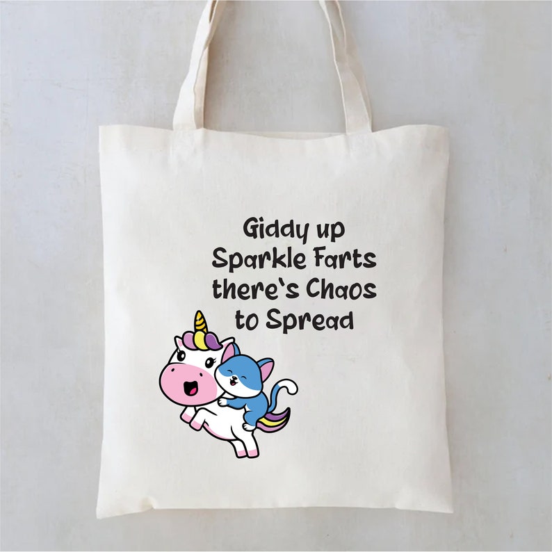 May include: A white tote bag with a black and white cartoon unicorn and a blue cat. The text on the bag reads "Giddy up Sparkle Farts there's Chaos to Spread".