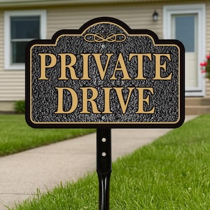 May include: A black and gold metal sign with the words "PRIVATE DRIVE" in gold lettering. The sign is mounted on a black metal post and is set in a grassy yard. The sign has a decorative top.