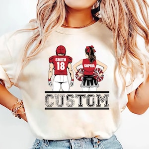 May include: Cream-colored t-shirt featuring a football player and a cheerleader. The football player wears a red helmet and jersey with the name "Smith" and the number "18." The cheerleader wears a red top with the name "Sophia" and holds pom-poms. The word "CUSTOM" is printed below.