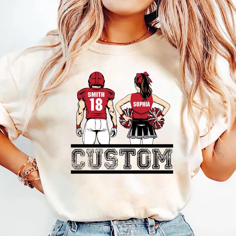 May include: Cream-colored t-shirt featuring a football player and a cheerleader. The football player wears a red helmet and jersey with the name "Smith" and the number "18." The cheerleader wears a red top with the name "Sophia" and holds pom-poms. The word "CUSTOM" is printed below.