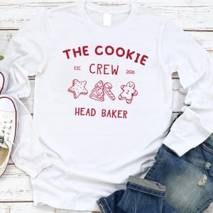 May include: A white long-sleeve shirt featuring red text that reads THE COOKIE CREW EST 2026 and HEAD BAKER. The design includes red line art illustrations of a star-shaped cookie, a Christmas tree, a candy cane, and a gingerbread man.