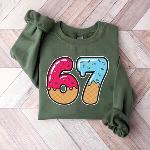 May include: An olive green sweatshirt featuring a colorful graphic of the number "67". The "6" is designed as a pink donut with sprinkles, and the "7" is blue with sprinkles, both on a waffle cone base.