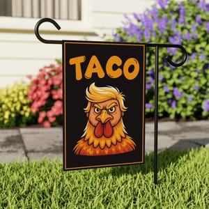 May include: A black garden flag with the word "TACO" in orange block letters. Below the text is a cartoon rooster with blonde hair and an angry expression. The flag is displayed on a black metal stand in a grassy yard.