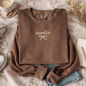 May include: A brown sweatshirt with the word "auntie" embroidered in white script, with a bow detail. A gold chain necklace is draped around the neckline. The sweatshirt is folded on top of blue denim shorts.
