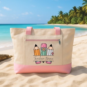 May include: Beige tote bag with pink accents, featuring a zippered pocket and pink handles. The front displays a graphic of pencils and a flower, with the text "Teacher Name" below. Perfect for carrying school supplies.