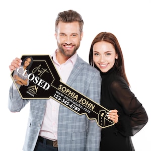 May include: A large black and gold key-shaped sign held by a smiling couple. The sign features the words "Just Closed", a realtor's name, website, and phone number, and an image of a realtor.