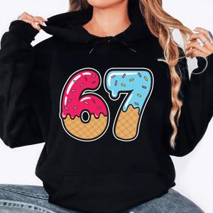 May include: Black hooded sweatshirt with a colorful graphic of the number 67. The number 6 is designed as a pink donut with sprinkles, and the number 7 is blue with sprinkles, resembling melting ice cream. The sweatshirt is made of a soft material.