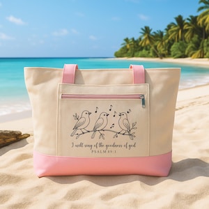 May include: A beige canvas tote bag with pink accents, featuring a black illustration of three singing birds on a branch. The bag has pink handles and a zippered pocket. Text on the bag reads "I will sing of the goodness of god, Psalm 89:1."