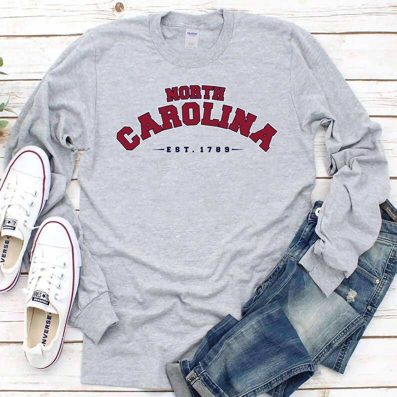 K&ouml;nnte beinhalten: Ein graues Langarmshirt mit einer roten und blauen Grafik, auf der vorne "NORTH CAROLINA - EST. 1789" steht. Das Shirt liegt auf einer Holzoberfl&auml;che mit einem Paar wei&szlig;en Turnschuhen und einer blauen Jeans.