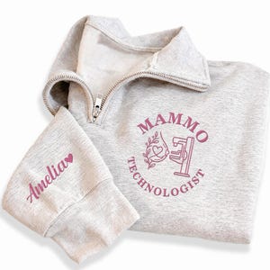 May include: A light gray quarter-zip sweatshirt featuring a pink graphic on the chest that reads Mammo Technologist with an illustration of a mammography machine and a heart. The sleeve is embroidered with the name Amelia and a small heart.