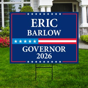 May include: A blue campaign sign with the name "ERIC BARLOW" and the title "GOVERNOR 2026" in white text. The sign has a red border and a section with blue stars and red and white stripes. It is placed on a green lawn.