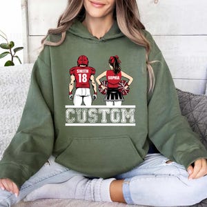 May include: Olive green hoodie featuring a custom design of a football player and cheerleader. The football player wears a red jersey with the name "Smith" and the number "18." The cheerleader is in a red top and black skirt, with the name "Sophia." The word "CUSTOM" is printed below.