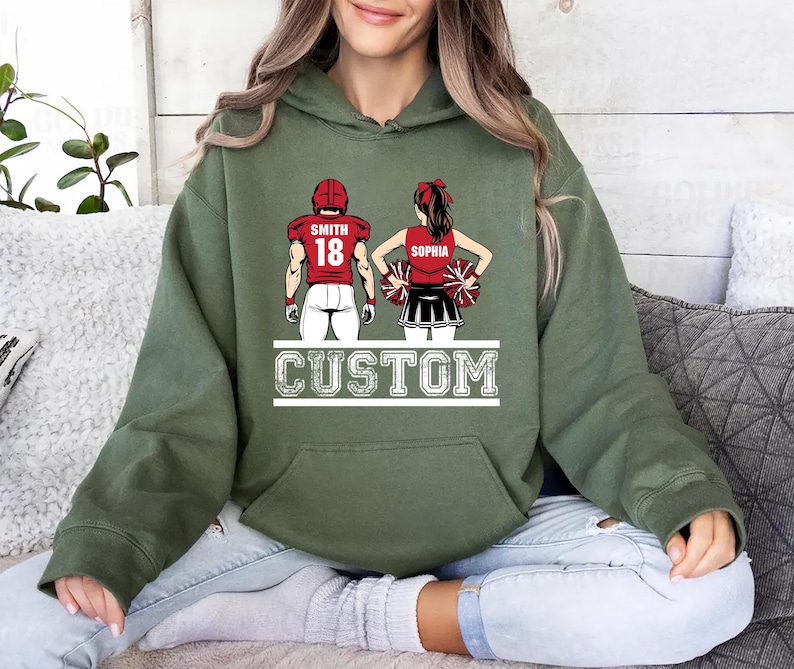 May include: Olive green hoodie featuring a custom design of a football player and cheerleader. The football player wears a red jersey with the name "Smith" and the number "18." The cheerleader is in a red top and black skirt, with the name "Sophia." The word "CUSTOM" is printed below.