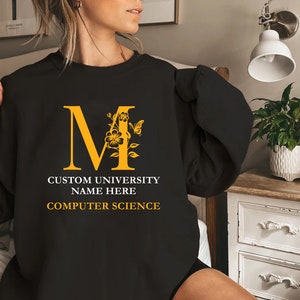 Peut inclure: Sweat-shirt noir avec une lettre M jaune décorée de fleurs et d'un papillon. Le texte "Custom University Name Here Computer Science" est imprimé sous la lettre.