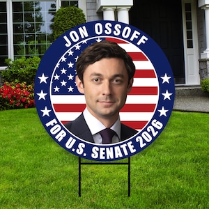 May include: A round political yard sign featuring a portrait of Jon Ossoff. The sign has a blue border with white stars and the text "JON OSSOFF" and "FOR U.S. SENATE 2026". The background is the American flag.