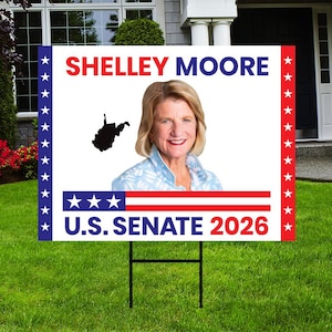 May include: A white yard sign with the text "SHELLEY MOORE" in red, a photo of a woman, and the text "U.S. SENATE 2026". The sign has a black metal stake and is on a green lawn.
