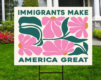 May include: A cream-colored yard sign with the text "IMMIGRANTS MAKE AMERICA GREAT" in teal. The sign features pink flowers with orange centers and teal leaves. The sign is displayed on a black metal stake in a grassy yard.