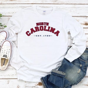 K&ouml;nnte beinhalten: Wei&szlig;es Langarm-T-Shirt mit einer roten und blauen Grafik, auf der "NORTH CAROLINA -EST. 1789- " steht. Das Shirt liegt flach auf einer wei&szlig;en Holzoberfl&auml;che mit einem Paar wei&szlig;en Turnschuhen und einer blauen Jeans.