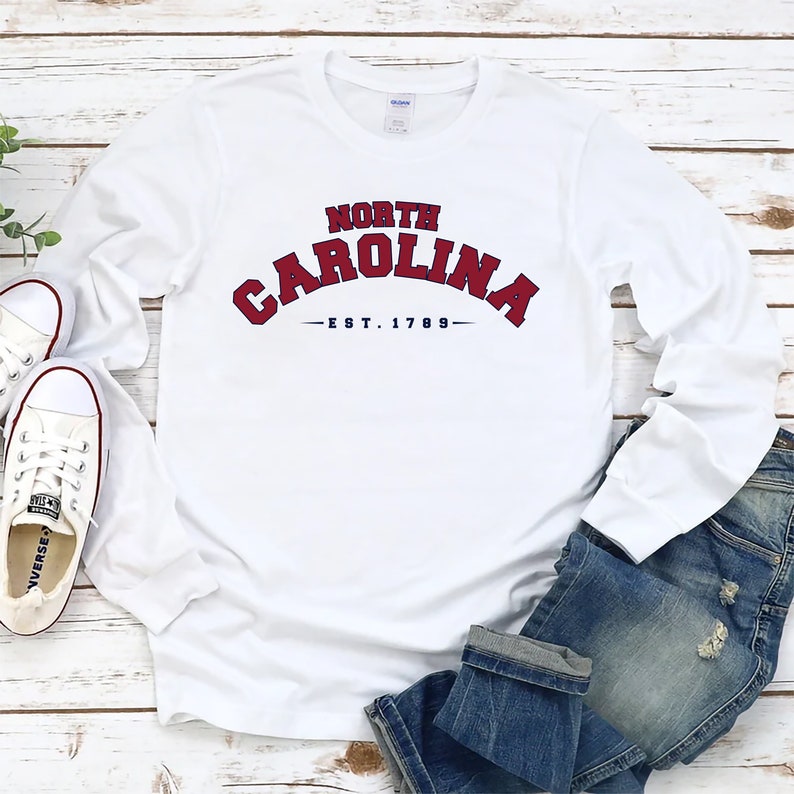 K&ouml;nnte beinhalten: Wei&szlig;es Langarm-T-Shirt mit einer roten und blauen Grafik, auf der "NORTH CAROLINA -EST. 1789- " steht. Das Shirt liegt flach auf einer wei&szlig;en Holzoberfl&auml;che mit einem Paar wei&szlig;en Turnschuhen und einer blauen Jeans.