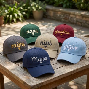 May include: Six baseball caps in various colors including dark gray, green, beige, maroon, navy blue, and light blue. Each cap features embroidered cursive text reading mimi, nana, nini, grandma, Mama, and gigi, with a small heart icon below.