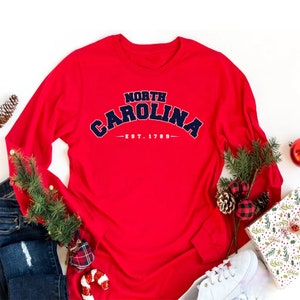 K&ouml;nnte beinhalten: Rotes Langarm-T-Shirt mit einer marineblauen Grafik, auf der "NORTH CAROLINA - EST. 1789" steht