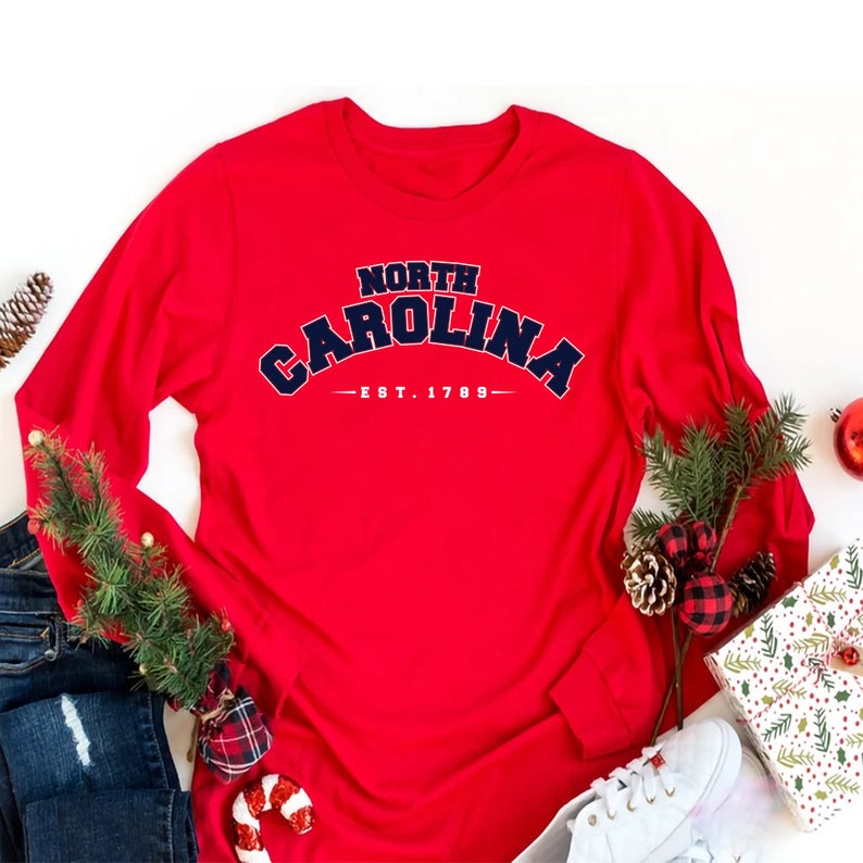 K&ouml;nnte beinhalten: Rotes Langarm-T-Shirt mit einer marineblauen Grafik, auf der "NORTH CAROLINA - EST. 1789" steht