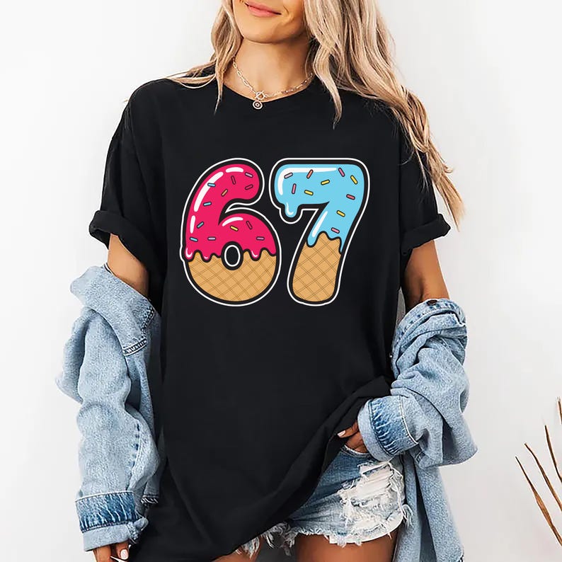 May include: Black t-shirt featuring a colorful graphic of the number 67 designed to look like a donut and ice cream cone. The number 6 is pink with sprinkles, and the 7 is blue with sprinkles. The shirt is worn by a person.
