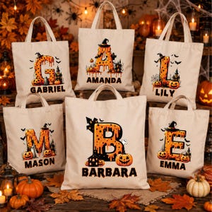 May include: A collection of six off-white canvas tote bags, each featuring a large orange letter with a dripping effect, a black witch hat, pumpkins, bats, and a haunted house silhouette, with a personalized name printed below each letter.