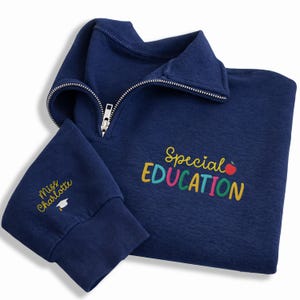May include: Navy blue quarter-zip sweatshirt with the words "Special EDUCATION" in colorful embroidery. The left sleeve cuff features the name "Miss Charlotte" and a graduation cap. The sweatshirt has a silver zipper.