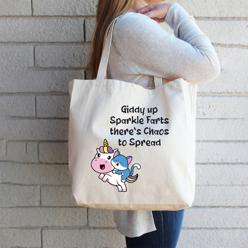 May include: A white canvas tote bag with a unicorn and cat graphic and the text "Giddy up Sparkle Farts there's Chaos to Spread".