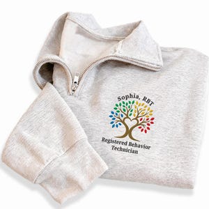 May include: A light gray quarter-zip sweatshirt featuring an embroidered tree design with multicolored leaves and a heart-shaped trunk. The text reads Sophia, RBT and Registered Behavior Technician in black lettering below the colorful tree graphic.