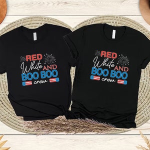 May include: Two black t-shirts with the text "Red White and Boo Boo Crew" in red, white, and blue. The shirts are on a woven wicker surface.