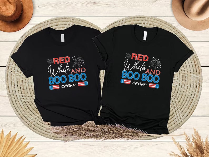 May include: Two black t-shirts with the text "Red White and Boo Boo Crew" in red, white, and blue. The shirts are on a woven wicker surface.