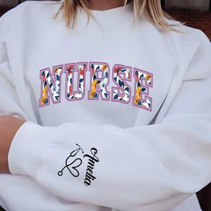 May include: White sweatshirt with the word "NURSE" printed in pink with floral accents. The sleeve has a stethoscope and heart graphic with the name "Amelia" written underneath.