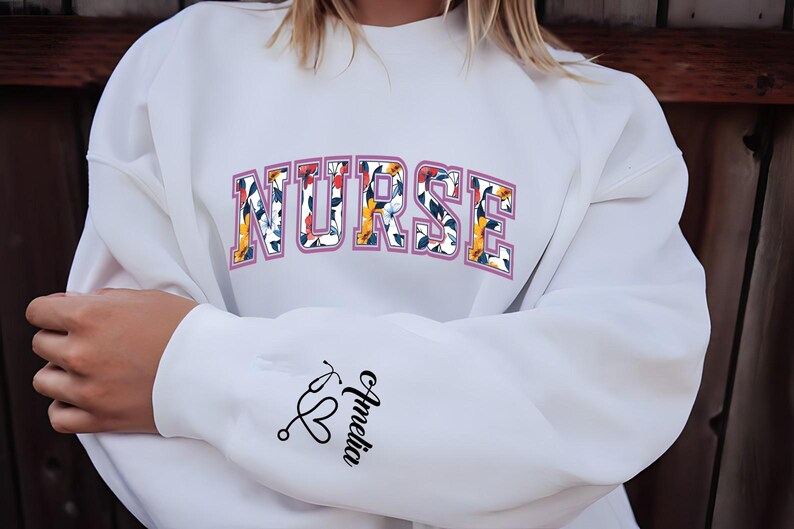 May include: White sweatshirt with the word "NURSE" printed in pink with floral accents. The sleeve has a stethoscope and heart graphic with the name "Amelia" written underneath.