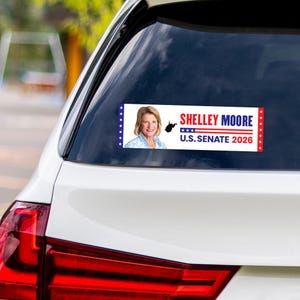 May include: A white car with a bumper sticker that reads "SHELLEY MOORE U.S. SENATE 2026." The sticker features a photo of a woman, the West Virginia state outline, and red, white, and blue stars and stripes.