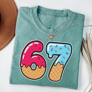 May include: A teal-colored t-shirt with a large graphic of the number 67. The number 6 is pink with sprinkles and a waffle cone base. The number 7 is blue with sprinkles and a waffle cone base. A gold necklace is draped across the shirt.