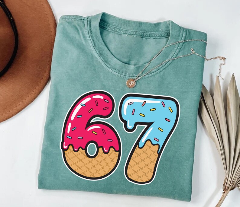 May include: A teal-colored t-shirt with a large graphic of the number 67. The number 6 is pink with sprinkles and a waffle cone base. The number 7 is blue with sprinkles and a waffle cone base. A gold necklace is draped across the shirt.