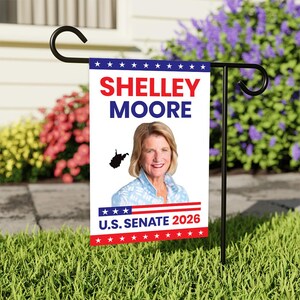 May include: A garden flag with the name "Shelley Moore" in red and blue, a photo of a woman, and the text "U.S. Senate 2026." The flag is on a black metal stand in a garden with green grass and colourful flowers.