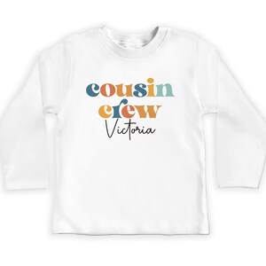 May include: White long-sleeve shirt with the text "cousin crew Victoria" in colorful retro-style lettering.