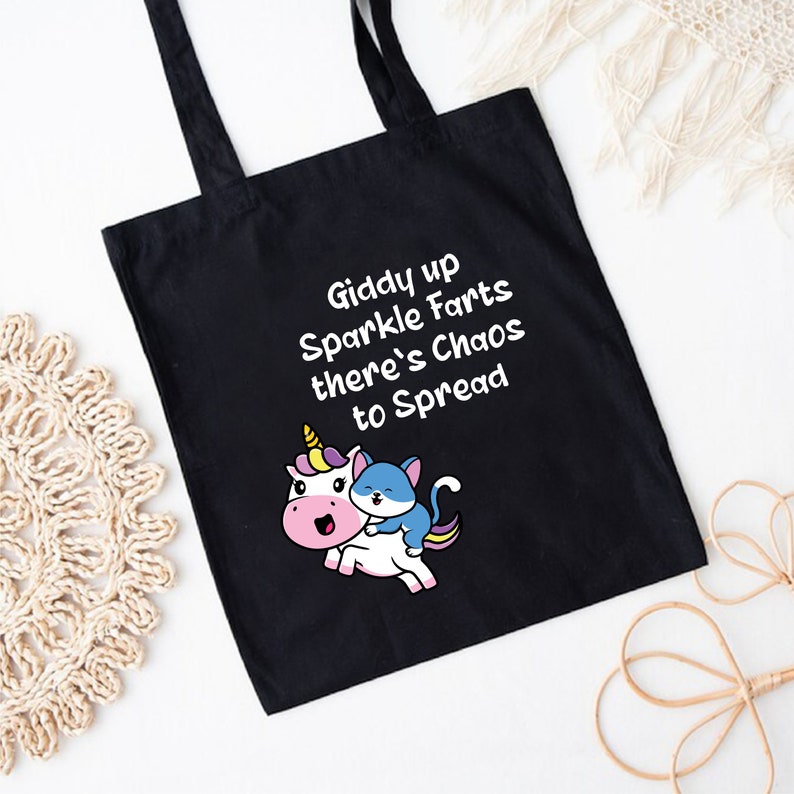 May include: Black tote bag with white text that reads "Giddy up Sparkle Farts there's Chaos to Spread". A cartoon unicorn with a blue cat riding on its back is printed on the bag.