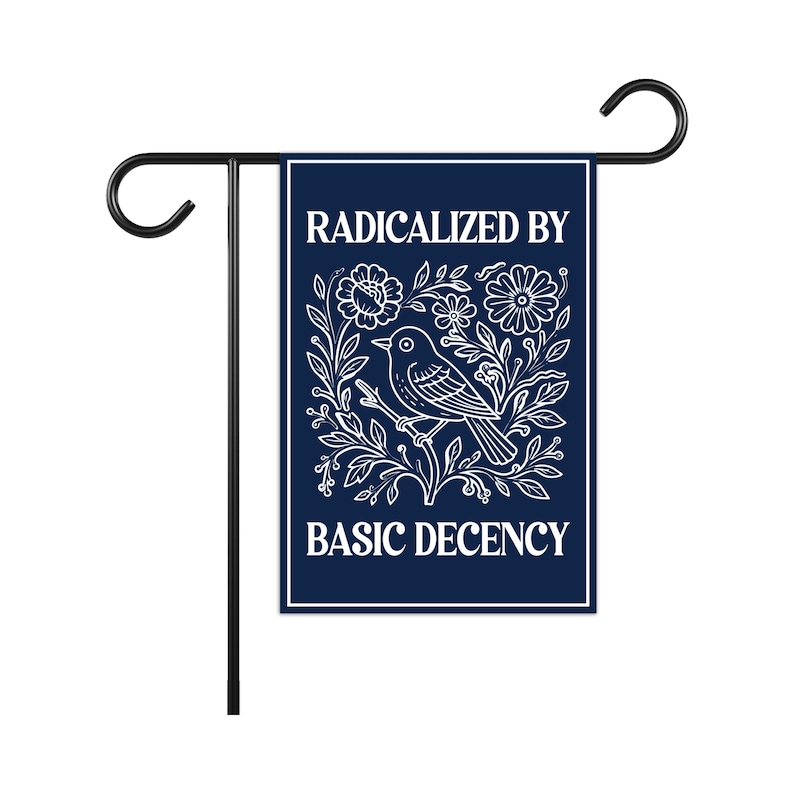 Puede incluir: Una bandera de jard&iacute;n azul marino con el texto "RADICALIZED BY BASIC DECENCY" en blanco. La bandera presenta una ilustraci&oacute;n blanca de un p&aacute;jaro en una rama rodeada de flores. Est&aacute; sujeta a una estaca de jard&iacute;n de metal negro.