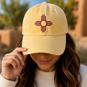 May include: A pale yellow baseball cap featuring a dark red embroidered Zia sun symbol centered on the front panel. The cap has a curved brim, a structured crown with a top button, and stitched eyelets for ventilation on each of the six panels.