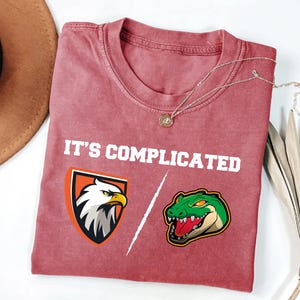 May include: A red t-shirt with the text "It's complicated" printed on it. The shirt features a graphic of an eagle and a crocodile on either side of the text.