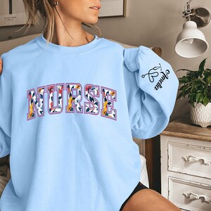 May include: Light blue sweatshirt with the word "NURSE" in floral print letters. The right sleeve has a small graphic of a stethoscope and the word "Nurse" written in cursive.