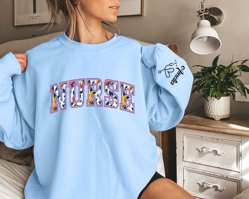 May include: Light blue sweatshirt with the word "NURSE" in floral print letters. The right sleeve has a small graphic of a stethoscope and the word "Nurse" written in cursive.
