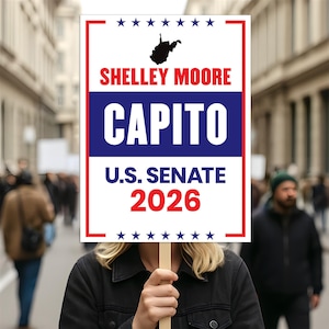 May include: A sign with the text "Shelley Moore Capito U.S. Senate 2026" in red and blue, with a black outline of West Virginia and stars. The sign is held by a person in front of a crowd of people.