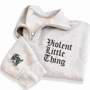 May include: A light gray sweatshirt with a quarter-zip collar. The text "Violent Little Thing" is printed in black on the front. A black dragon graphic is on the left sleeve. The sweatshirt is made of a soft material.