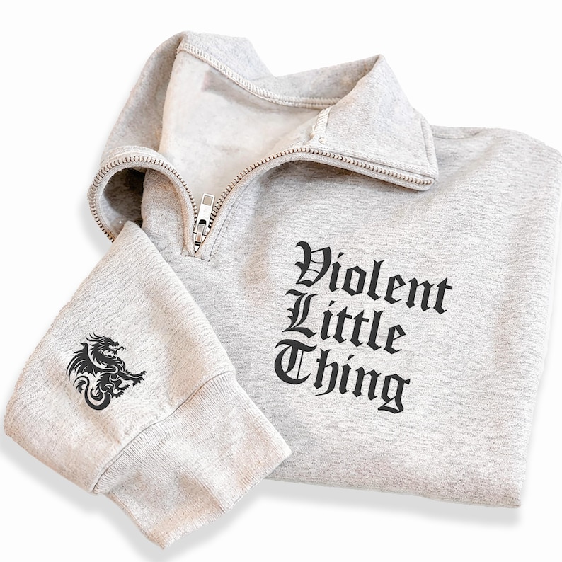 May include: A light gray sweatshirt with a quarter-zip collar. The text "Violent Little Thing" is printed in black on the front. A black dragon graphic is on the left sleeve. The sweatshirt is made of a soft material.