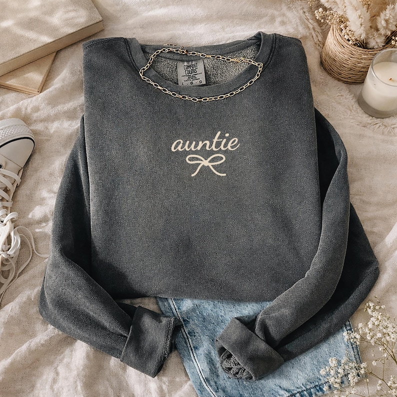 May include: A gray sweatshirt with the word "auntie" embroidered in white script, along with a bow design. A gold chain necklace rests on the collar. The sweatshirt is paired with denim shorts.